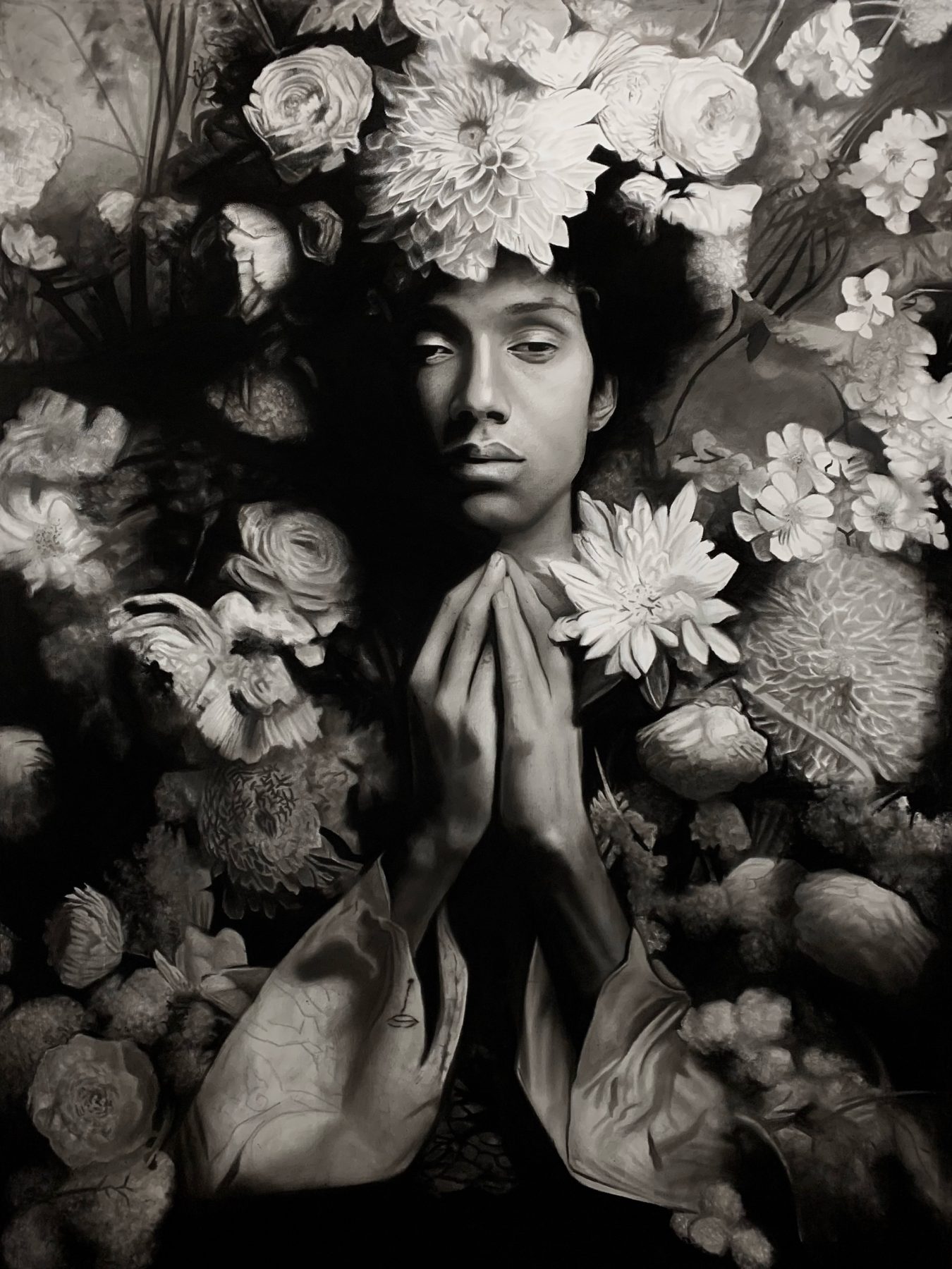 Jardim das fragilidades I by Ritchelly Oliveira: a young man with his hands pressed together in front of his chest, surrounded by dense flowers, in black and white