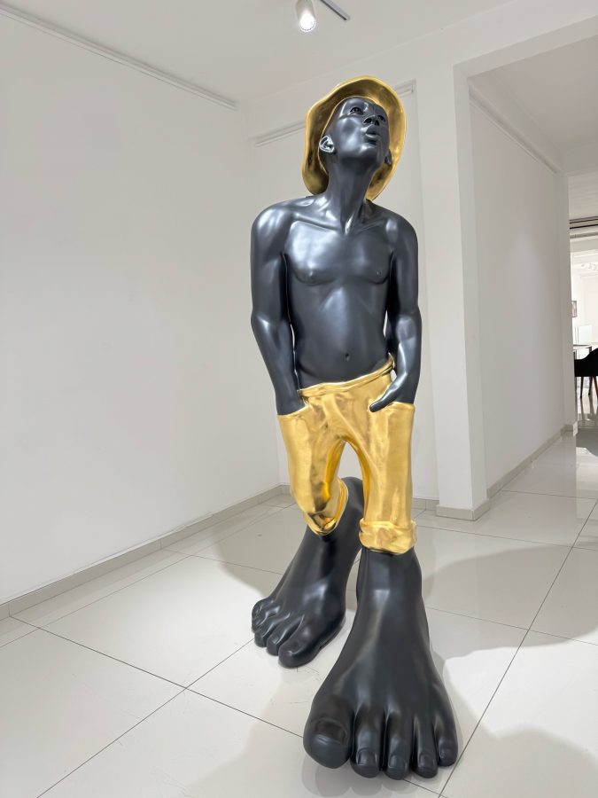 Le Siffleur 220 by Idan Zareski, monumental resin sculpture of a whistling man with a dark body, gold hat and gold trousers, hands in pockets, standing on enormous bare feet in the gallery