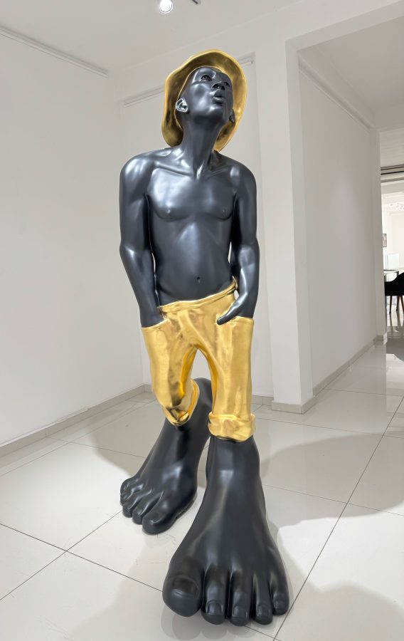 Le Siffleur 220 by Idan Zareski, monumental resin sculpture of a whistling man with a dark body, gold hat and gold trousers, hands in pockets, standing on enormous bare feet in the gallery